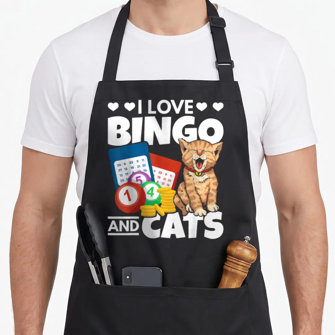 Cat bingo apron - Orange cat sitting next to blue and red bingo cards, colored balls, and gold coin stacks under white — shown on a black cotton canvas apron against a white background