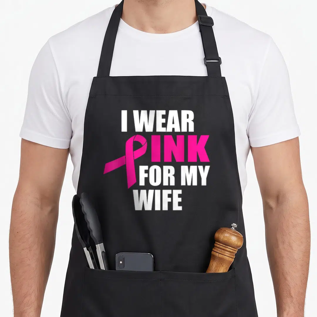 Cancer wife apron - White stacked text I WEAR PINK FOR MY WIFE with pink ribbon overlapping the letter P — shown on a black cotton canvas apron against a white background