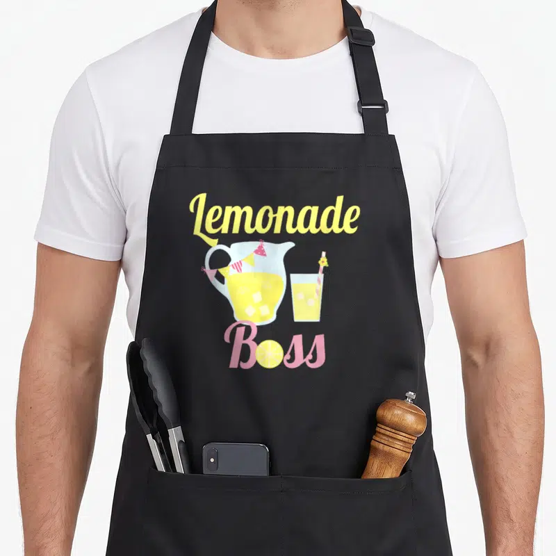 Stand boss lemonade apron - Lemonade Boss text with pitcher, glass, lemon slice o, and bunting flags in yellow pink — shown on a black cotton canvas apron against a white background