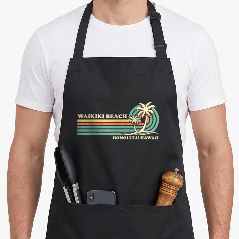 Hawaii summer aprons - Retro Waikiki Beach text with striped sun and two palm trees in teal and orange — shown on a black cotton canvas apron against a white background