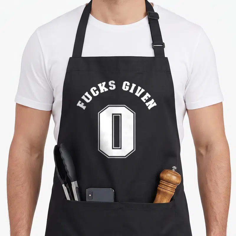 Profanity zero apron - White arched text reading Fucks Given 0 above a geometric number zero — shown on a black cotton canvas apron against a white background