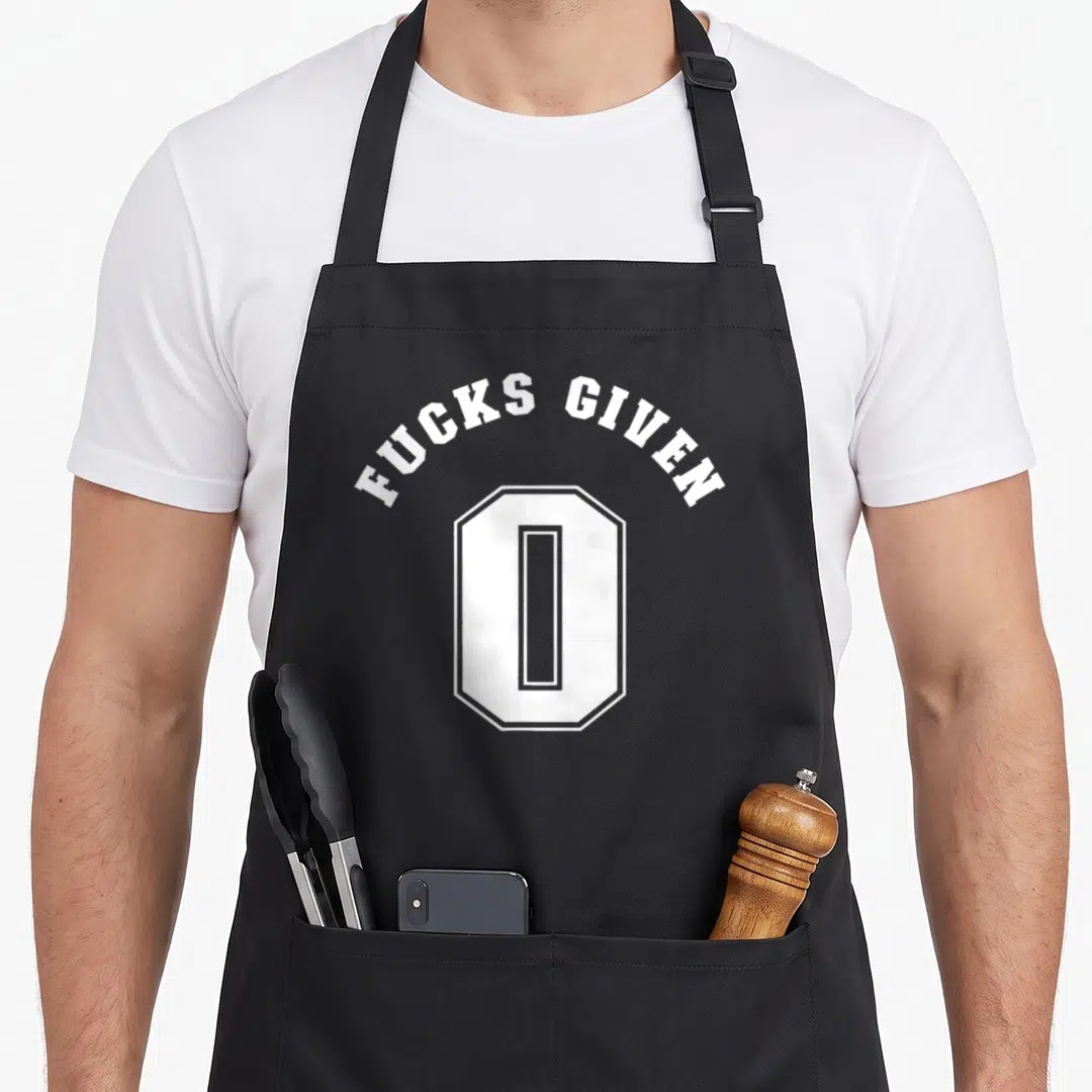 Profanity zero apron - White arched text reading Fucks Given 0 above a geometric number zero — shown on a black cotton canvas apron against a white background