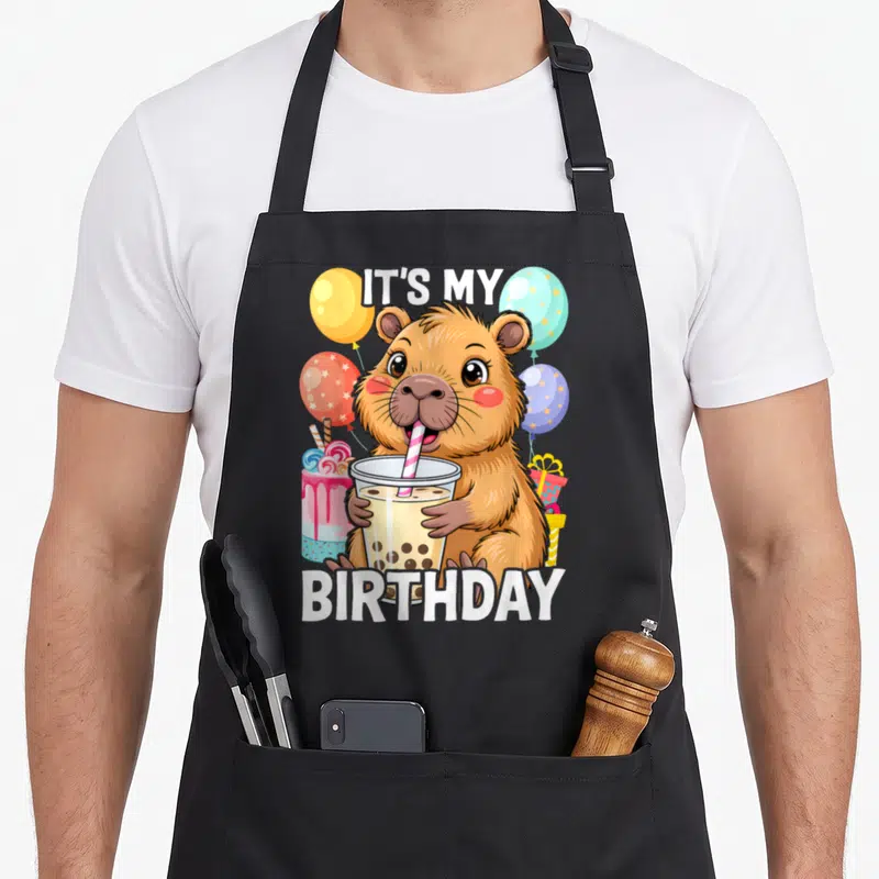 Capibara birthday apron - Cartoon capybara holding bubble tea with IT'S MY BIRTHDAY text and balloons — shown on a black cotton canvas apron against a white background