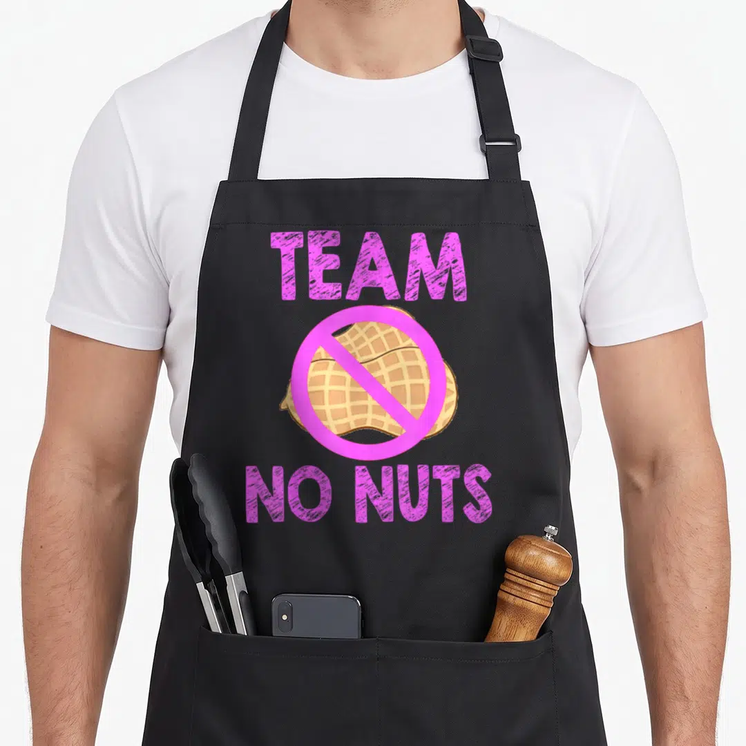 Nut side addict apron - TEAM NO NUTS text with pink prohibition sign over tan cartoon peanut — shown on a black cotton canvas apron against a white background