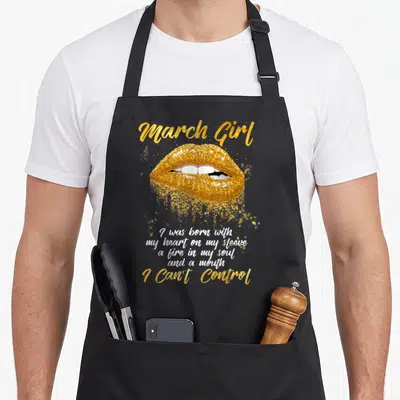 Funny march birthday apron - Gold glittery lips with white text reading March Girl and a mouth I Can't Control — shown on a black cotton canvas apron against a white background