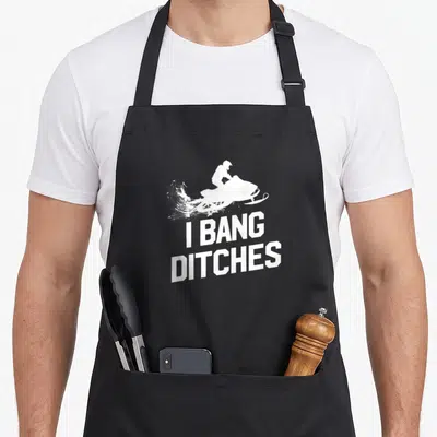 Bang snowmobile girl apron - White snowmobile rider silhouette above stacked I Bang Ditches text — shown on a black cotton canvas apron against a white background