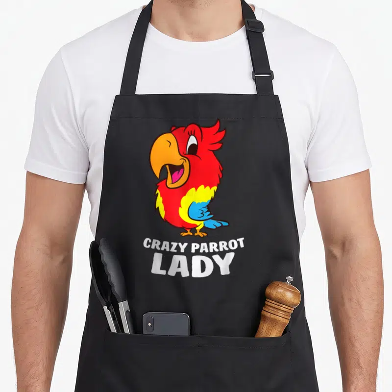 Vintage crazy parrot lady apron - Red cartoon parrot with yellow wing, blue tail, and white stacked text below — shown on a black cotton canvas apron against a white background
