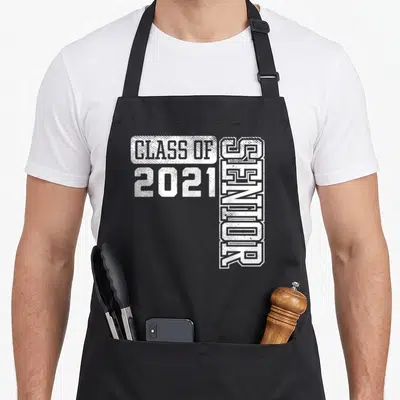 Class school graduation apron - White distressed text reading Class of 2021 Senior with vertical senior lettering — shown on a black cotton canvas apron against a white background