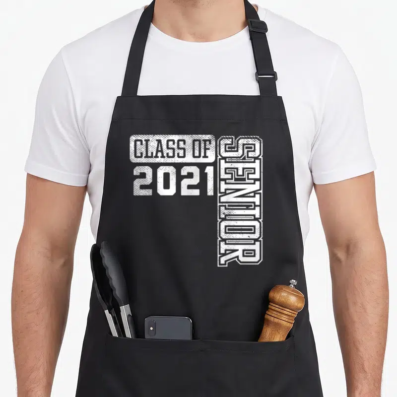 Class school graduation apron - White distressed text reading Class of 2021 Senior with vertical senior lettering — shown on a black cotton canvas apron against a white background
