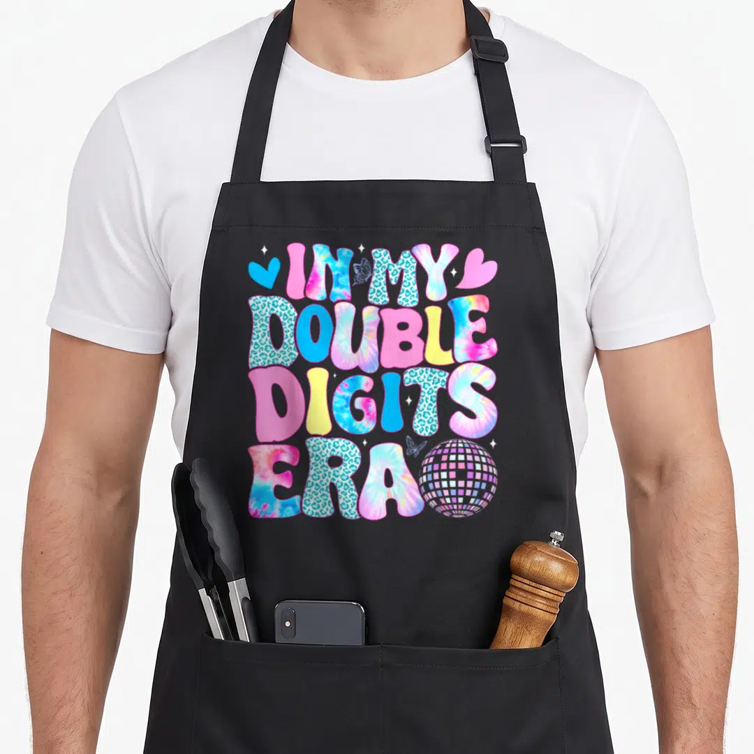 Digit birthday apron - Stacked text IN MY DOUBLE DIGITS ERA with tie-dye, leopard print, hearts, butterflies, and disco — shown on a black cotton canvas apron against a white background