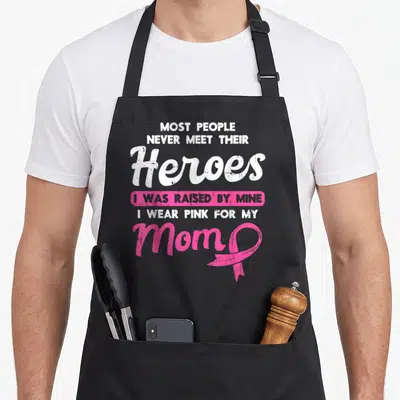 Raised breast cancer awareness aprons - Stacked text I Was Raised By Mine with pink ribbon next to Mom — shown on a black cotton canvas apron against a white background