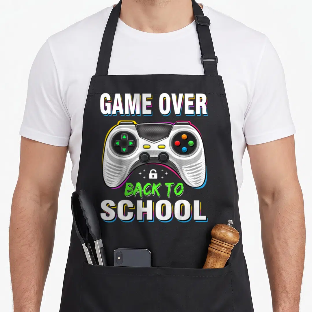 School game aprons - GAME OVER BACK TO SCHOOL text around a white and grey game controller with colored buttons — shown on a black cotton canvas apron against a white background