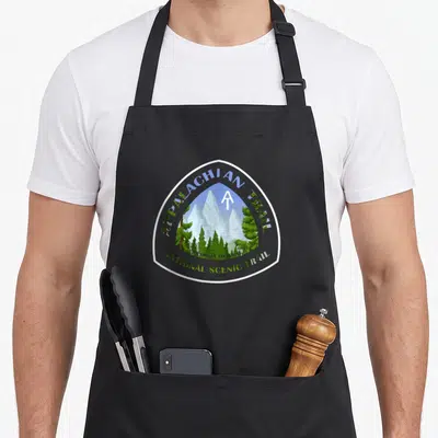 Scenic trailer girl apron - Shield emblem with mountains, trees, and Appalachian Trail text — shown on a black cotton canvas apron against a white background