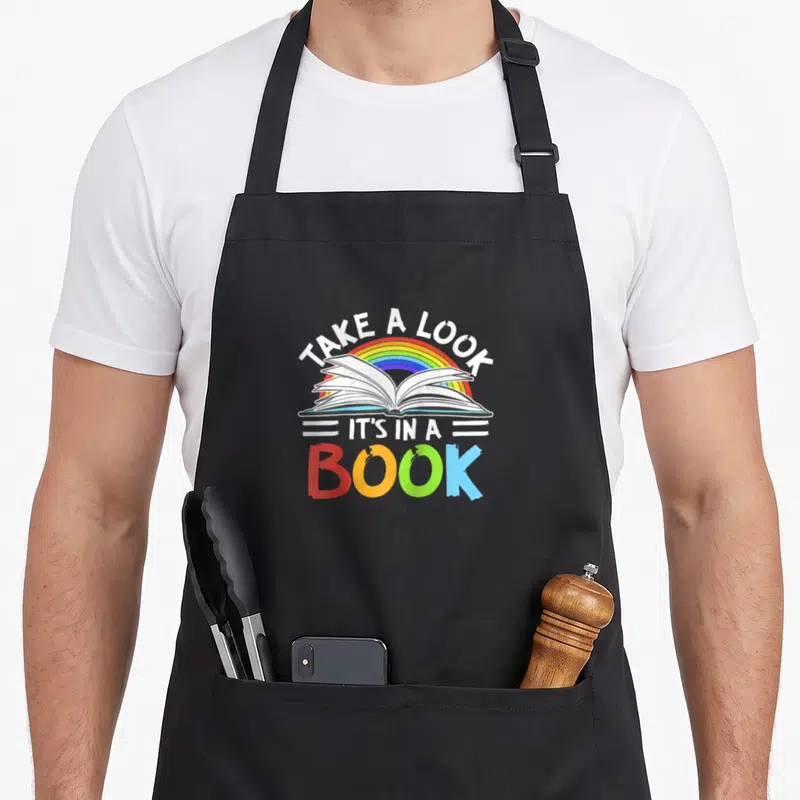 Baseball rainbow apron - Open book under rainbow arch with text TAKE A LOOK IT'S IN A BOOK in rainbow colors — shown on a black cotton canvas apron against a white background