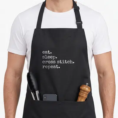 Eat cross stitch aprons - White stacked text reading eat sleep cross stitch repeat — shown on a black cotton canvas apron against a white background