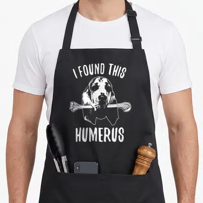 Humerus dog apron - White bloodhound holding a bone with text I FOUND THIS HUMERUS on grey — shown on a black cotton canvas apron against a white background