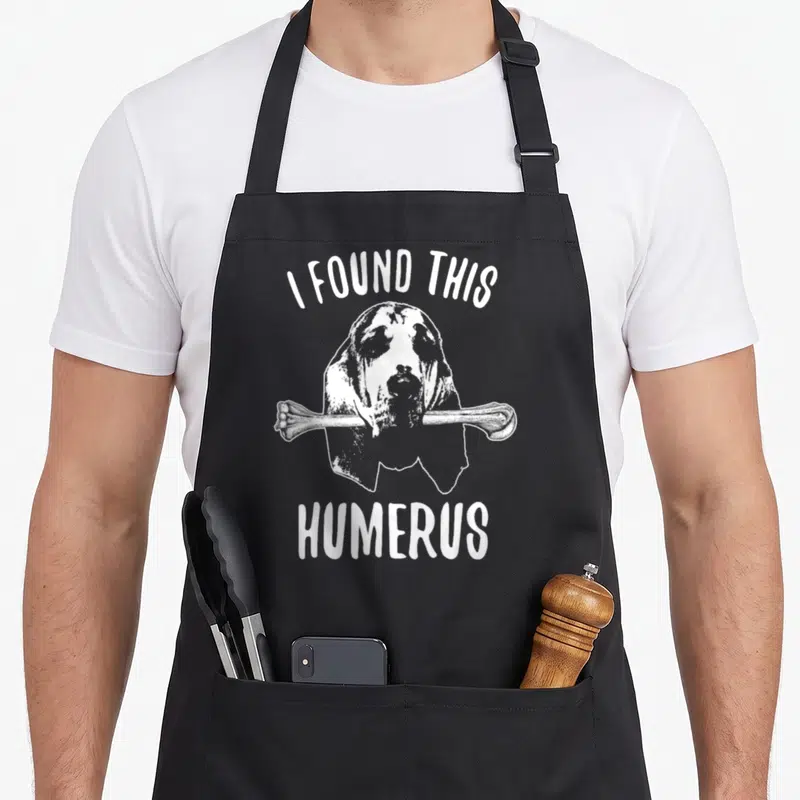 Humerus dog apron - White bloodhound holding a bone with text I FOUND THIS HUMERUS on grey — shown on a black cotton canvas apron against a white background