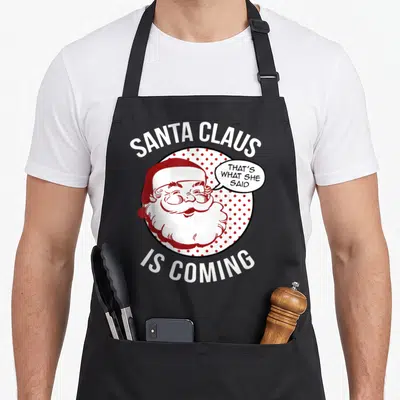 Coming santa claus apron - Santa Claus cartoon in red polka dot circle with speech bubble text — shown on a black cotton canvas apron against a white background