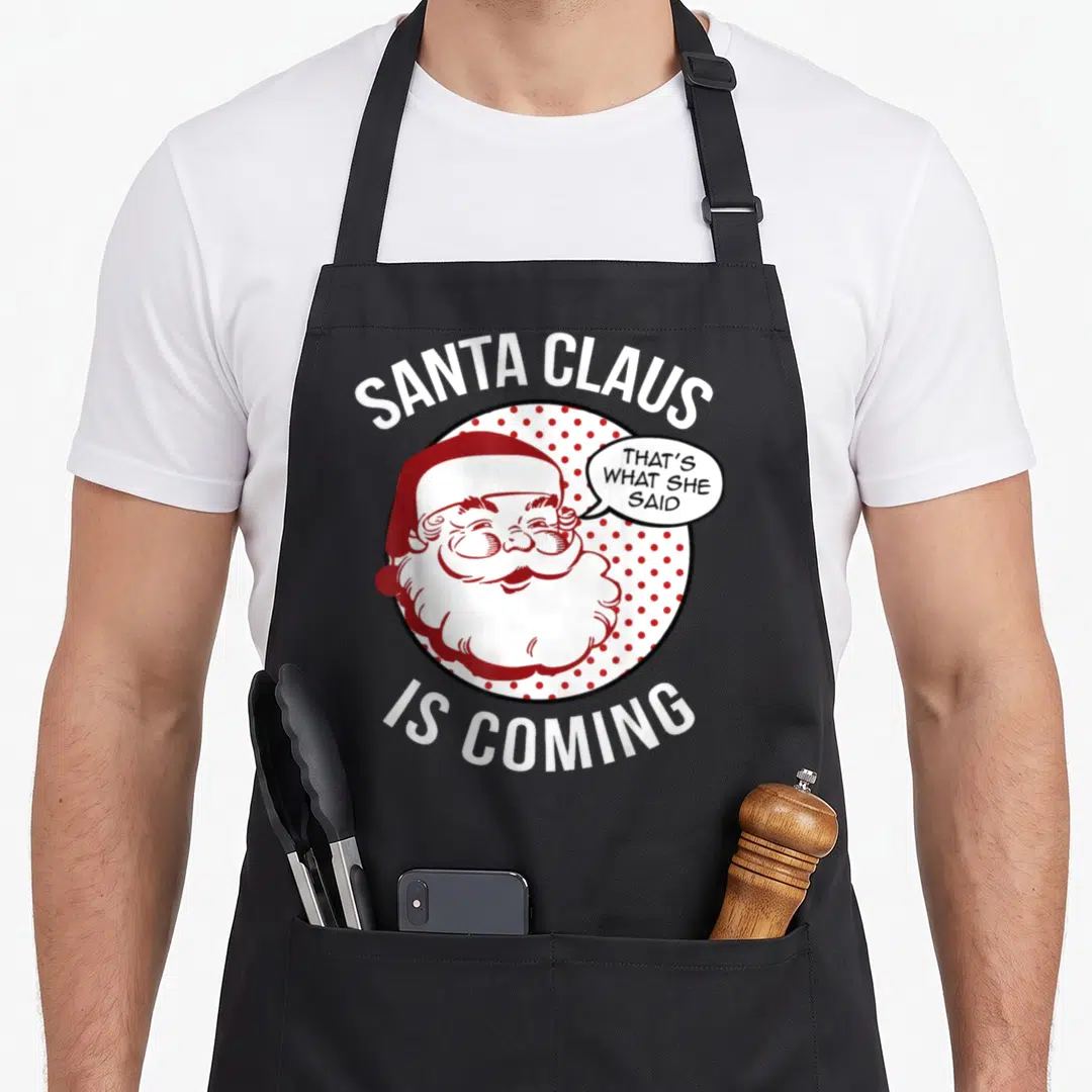 Coming santa claus apron - Santa Claus cartoon in red polka dot circle with speech bubble text — shown on a black cotton canvas apron against a white background