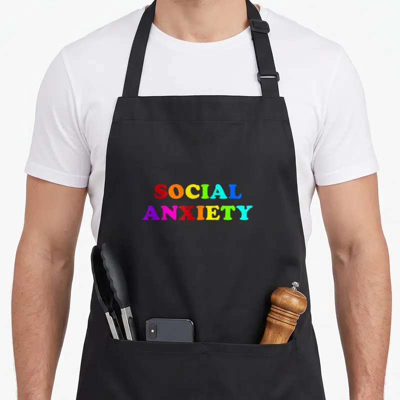 Alternative clothe goth apron - Stacked SOCIAL ANXIETY text in red orange yellow green blue purple pink teal — shown on a black cotton canvas apron against a white background