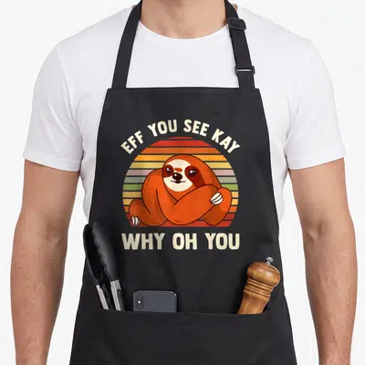 Sloth grill instructor apron - Cartoon sloth over striped sun with EFF YOU SEE KAY WHY OH YOU text — shown on a black cotton canvas apron against a white background