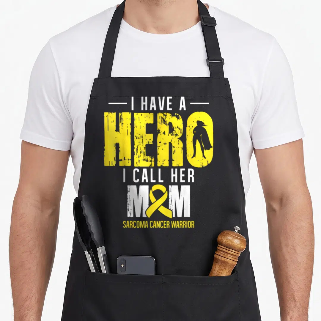 Call ribbon apron - Yellow ribbon and superhero silhouette inside text reading I Have A Hero I Call Her Mom Sarcoma — shown on a black cotton canvas apron against a white background