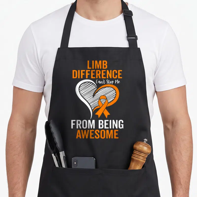 Difference ribbon apron - White heart with orange outline and ribbon, text Limb Difference Can't Stop Me From Being — shown on a black cotton canvas apron against a white background