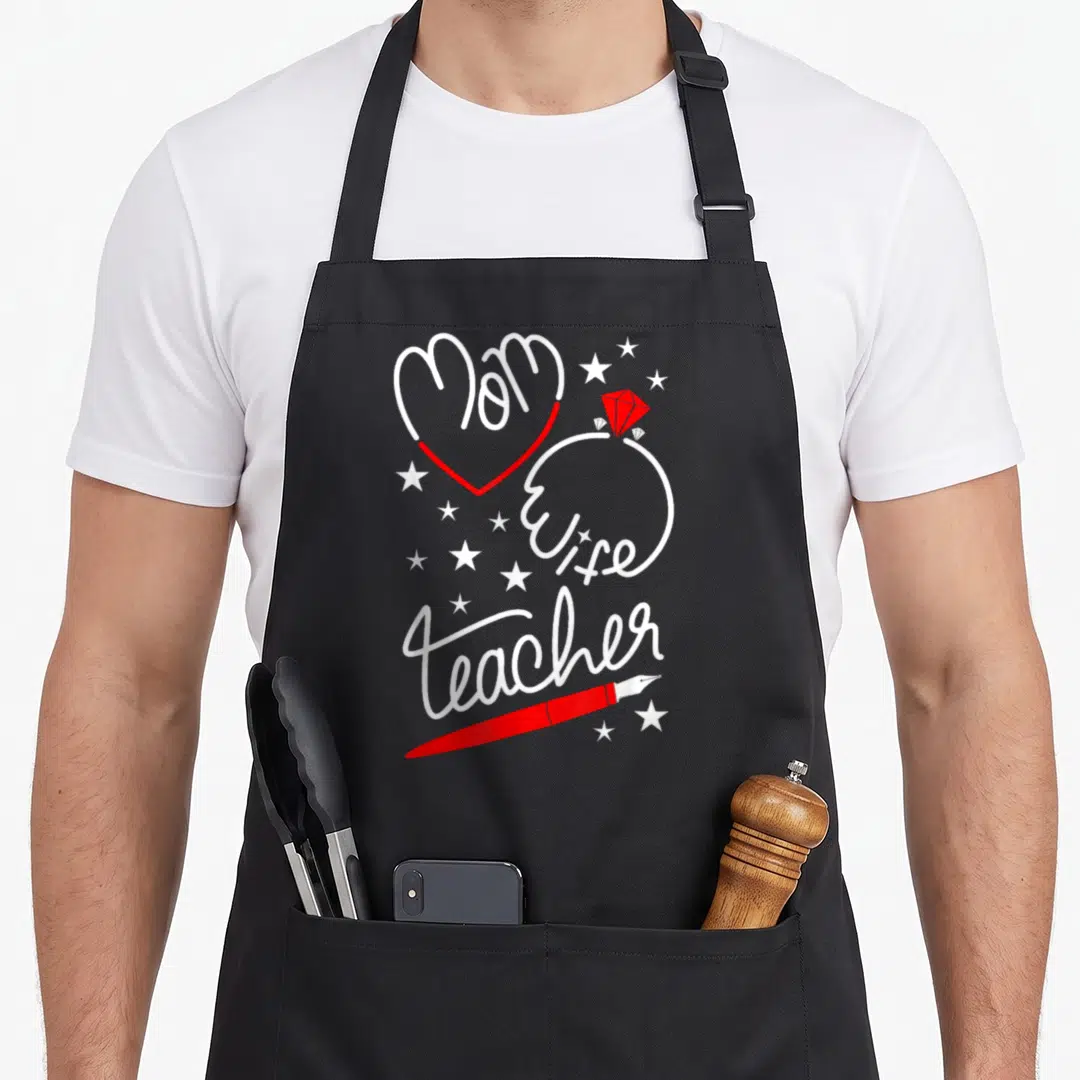 School best teacher apron - Stacked Mom Wife teacher text with red heart, diamond ring, red pen, and white stars — shown on a black cotton canvas apron against a white background