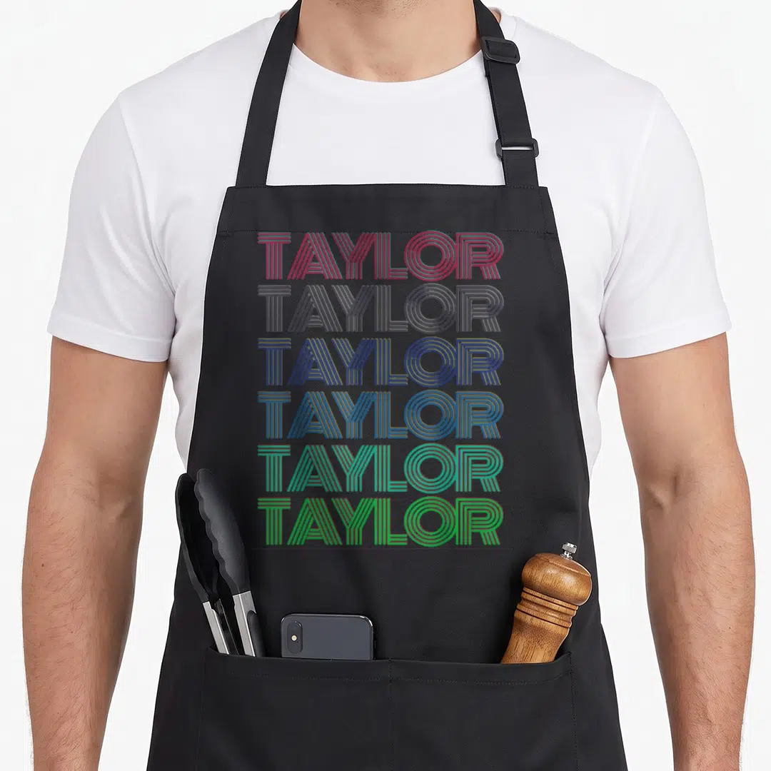 Cute taylor 80s apron - Stacked TAYLOR text in red grey blue teal and green lines — shown on a black cotton canvas apron against a white background