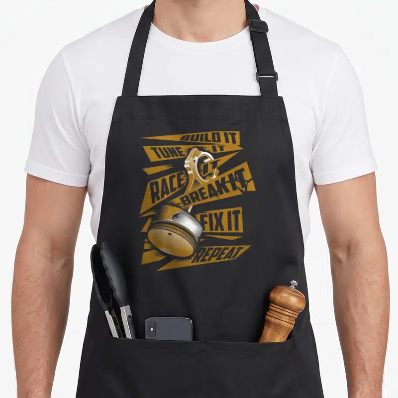 Tune air show apron - Engine piston and bearing with BUILD IT TUNE IT RACE IT BREAK IT FIX IT REPEAT text on yellow — shown on a black cotton canvas apron against a white background