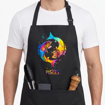 Pisce sign zodiac aprons - Two watercolor fish in a circle with PISCES text in blue yellow red purple green — shown on a black cotton canvas apron against a white background