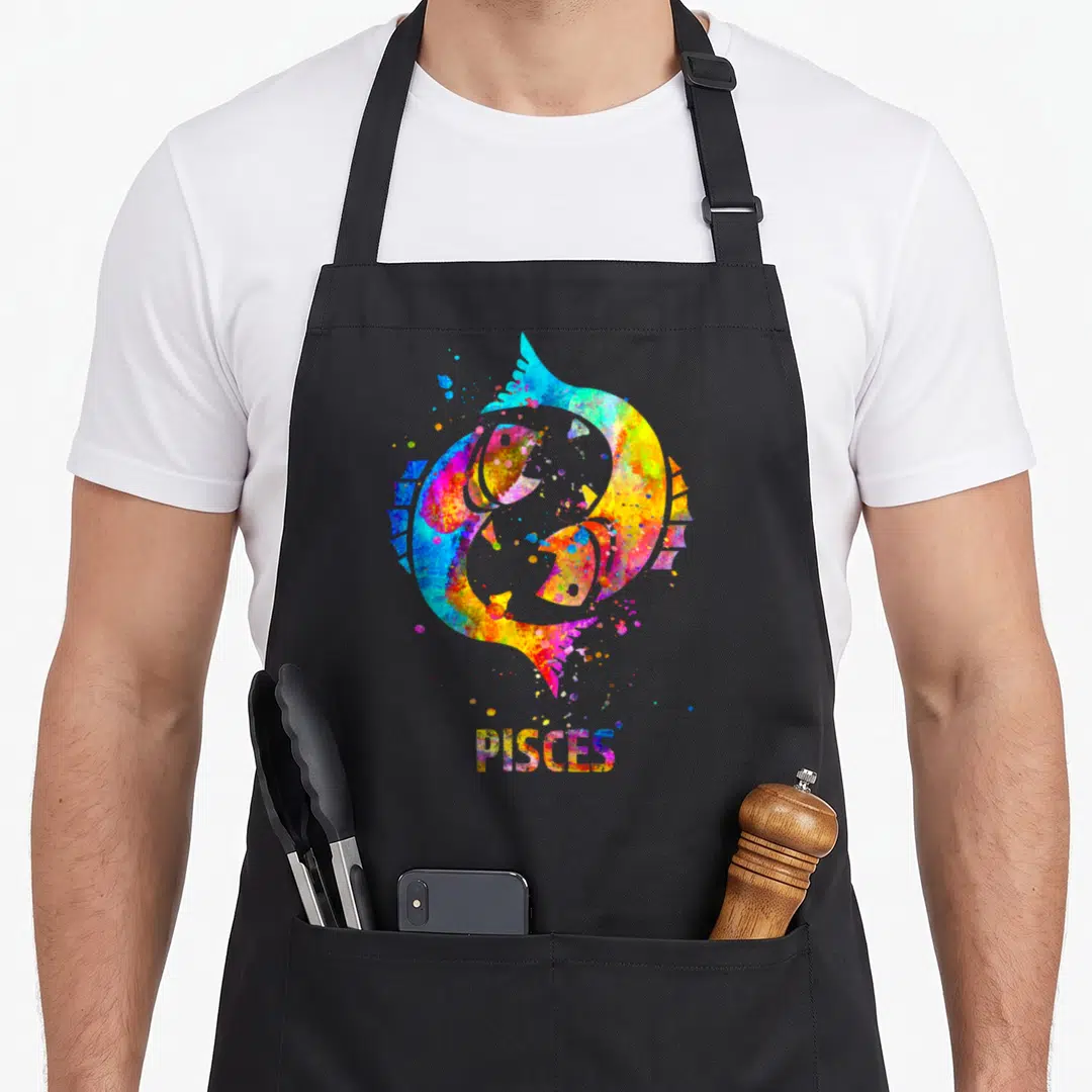 Pisce sign zodiac aprons - Two watercolor fish in a circle with PISCES text in blue yellow red purple green — shown on a black cotton canvas apron against a white background