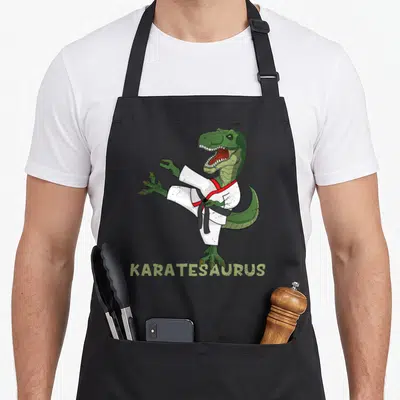 Karatesaurus dinosaur apron - Green cartoon dinosaur in white gi kicking with KARATESAURUS text below — shown on a black cotton canvas apron against a white background