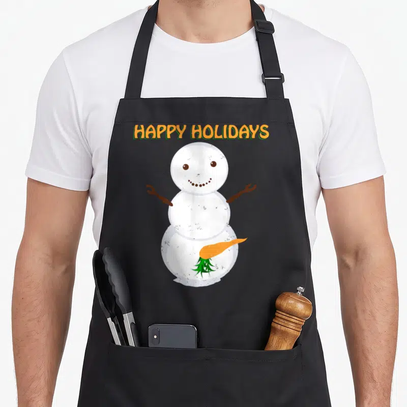 Jon ugly christmas apron - HAPPY HOLIDAYS text above a snowman with a fallen orange carrot lying on its base — shown on a black cotton canvas apron against a white background