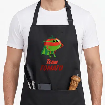 Tomato super hero aprons - Cartoon red and green tomato superhero standing above stacked text reading Team Tomato — shown on a black cotton canvas apron against a white background
