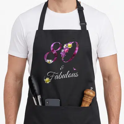 80th floral aprons - Number 80 filled with purple pink and yellow flowers above white script text and a single rose — shown on a black cotton canvas apron against a white background