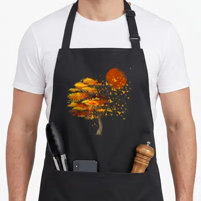 Bird tree apron - Realistic tree with autumn leaves, large sun, and birds in flight against grey background — shown on a black cotton canvas apron against a white background