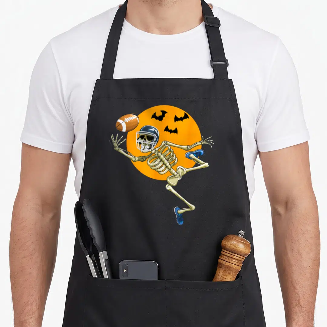 Football skeleton apron - Cartoon skeleton in helmet and blue shoes jumping to catch football against orange moon — shown on a black cotton canvas apron against a white background