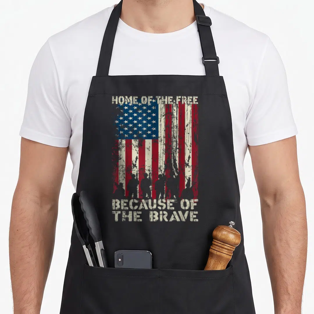 Brave american flag apron - Distressed USA flag with soldier silhouettes and Home of the Free Because of the Brave text — shown on a black cotton canvas apron against a white background