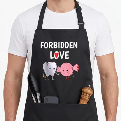 Forbidden dentist apron - FORBIDDEN LOVE text, broken heart O, crying tooth holding rose, holding hands with pink candy — shown on a black cotton canvas apron against a white background