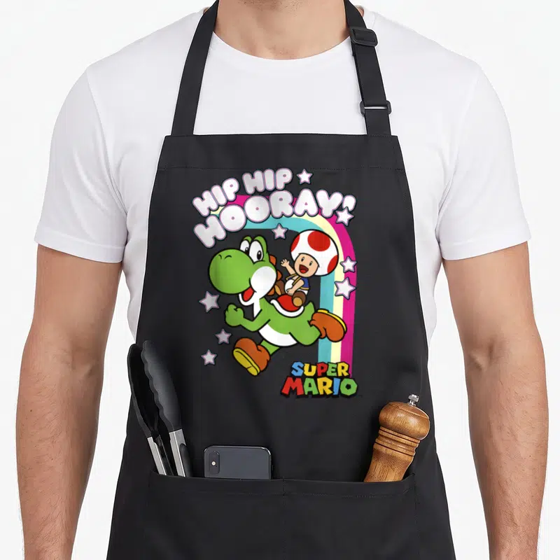 Toad rainbow apron - Yoshi carrying Toad under arched HIP HIP HOORAY! text and rainbow with scattered white stars — shown on a black cotton canvas apron against a white background