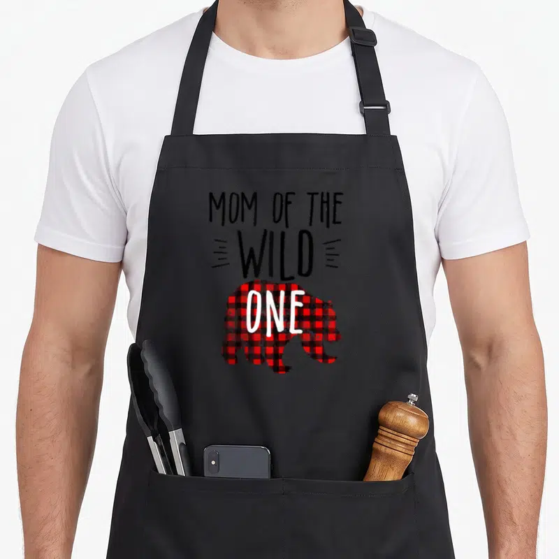 Wild buffalo plaid apron - MOM OF THE WILD ONE text above red and black checkered bear silhouette with ONE on its side — shown on a black cotton canvas apron against a white background