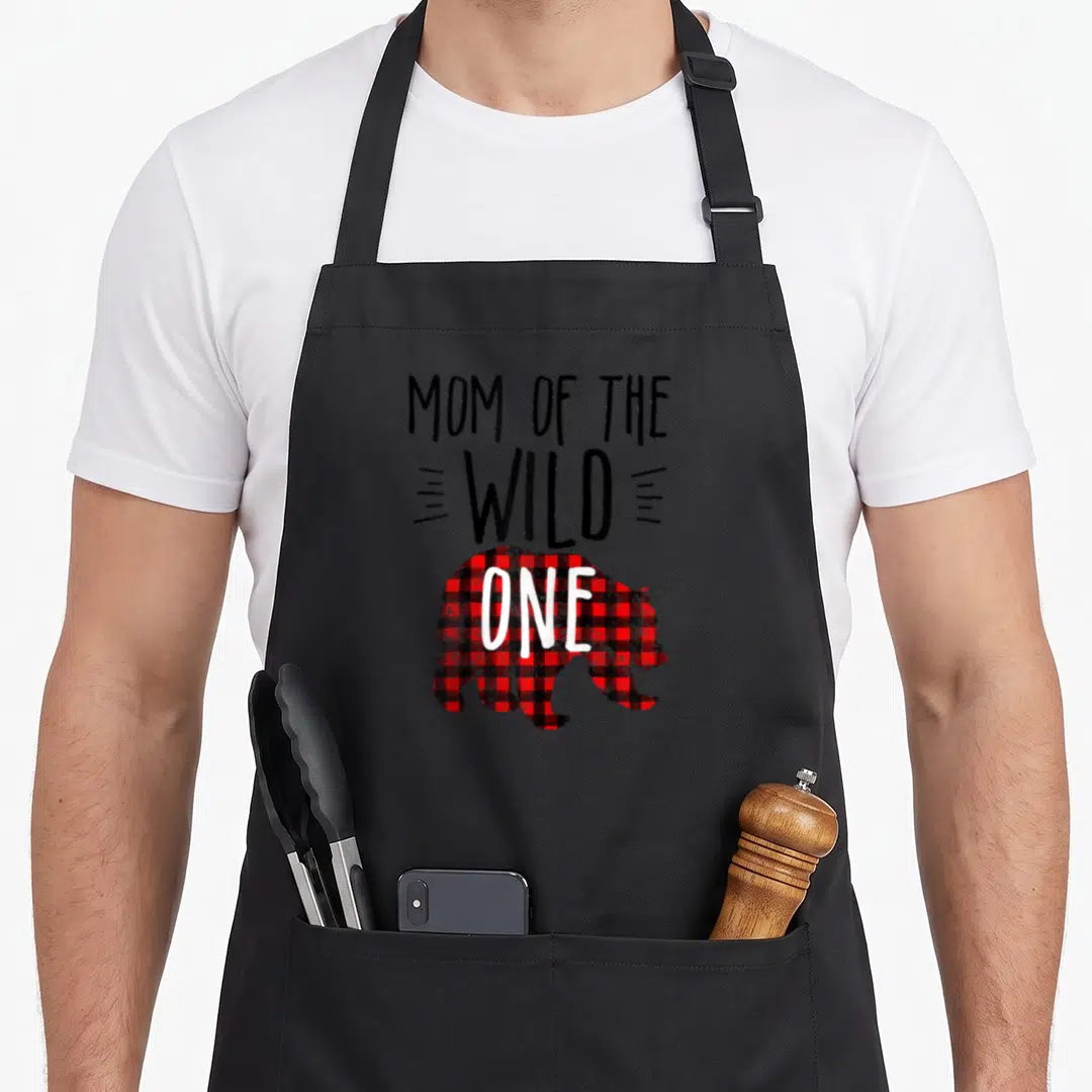 Wild buffalo plaid apron - MOM OF THE WILD ONE text above red and black checkered bear silhouette with ONE on its side — shown on a black cotton canvas apron against a white background