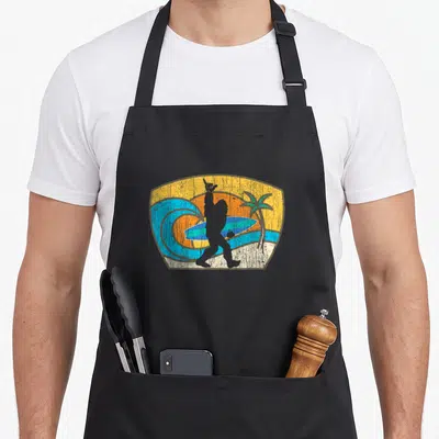 Bigfoot dude apron - Retro surfer silhouette holding board and shaka sign in shield with sun and palm tree — shown on a black cotton canvas apron against a white background