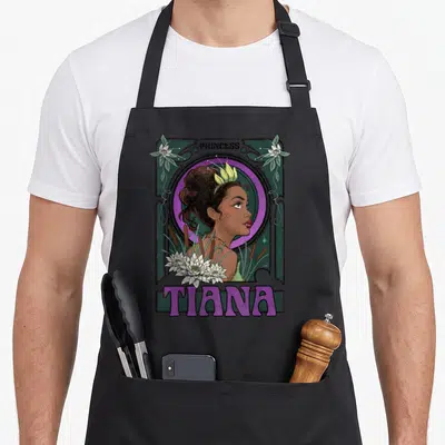 Profile princess apron - Tiana profile in purple circle with water lilies and cattails framed in green — shown on a black cotton canvas apron against a white background