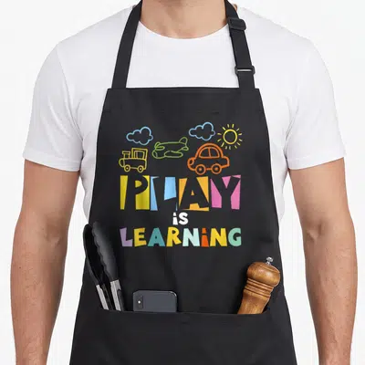 Play preschool teacher apron - Cartoon train, plane, car, sun, and clouds above multi-colored text reading PLAY is — shown on a black cotton canvas apron against a white background