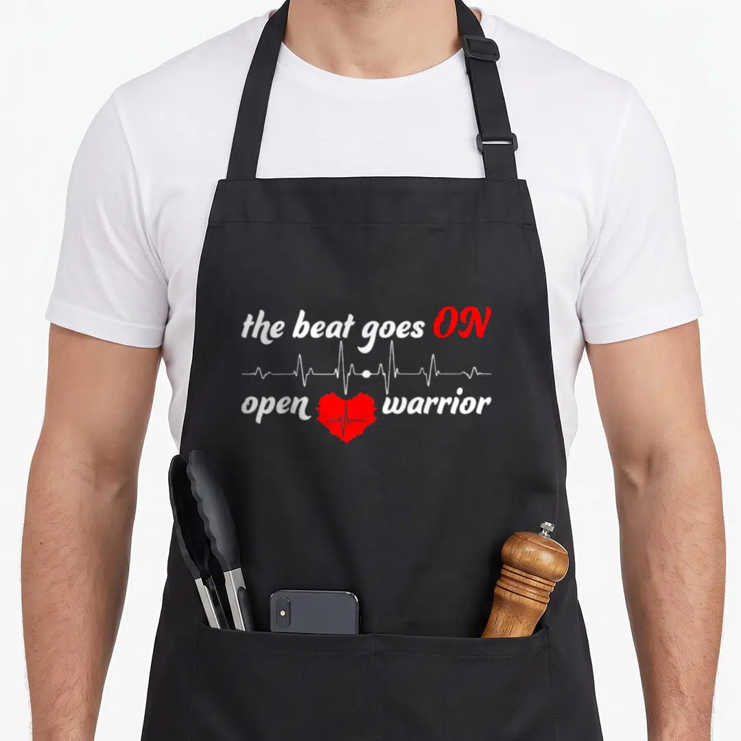 Post surgery open apron - White heartbeat line above red heart with pulse trace and open warrior text — shown on a black cotton canvas apron against a white background