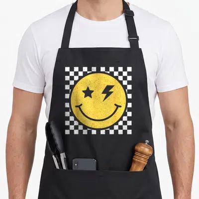 Pattern smile face on apron - Yellow smiley face with star and lightning eyes on white grey checkerboard — shown on a black cotton canvas apron against a white background