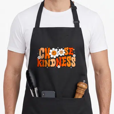 Choose unity day apron - Choose Kindness text with smiling daisy O's and stars in orange white and yellow cartoon style — shown on a black cotton canvas apron against a white background