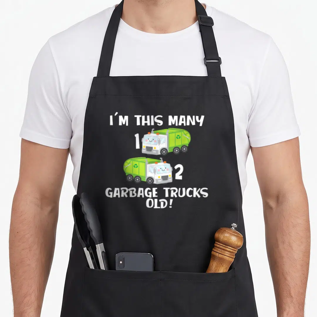 Toddle truck apron - Two green cartoon garbage trucks numbered one and two with white text reading I'M THIS MANY 1, 2 — shown on a black cotton canvas apron against a white background